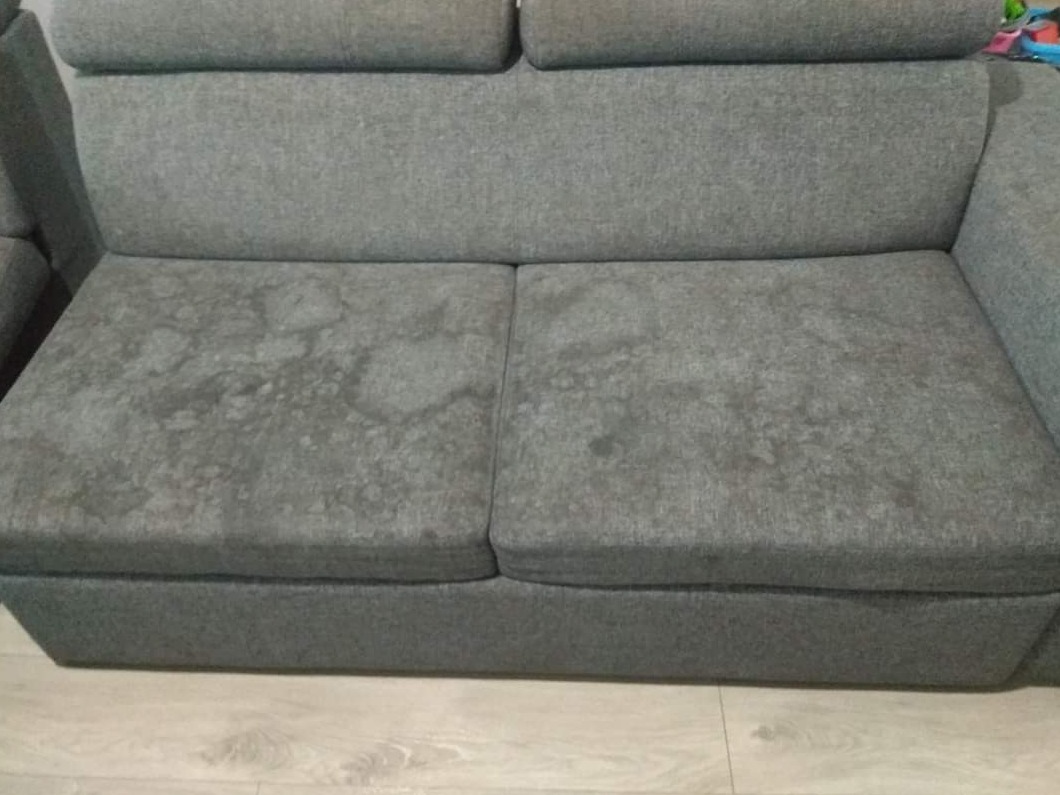 Szara, dwuosobowa kanapa z widocznymi, ciemniejszymi plamami na siedzisku, stojąca na podłodze imitującej drewno.