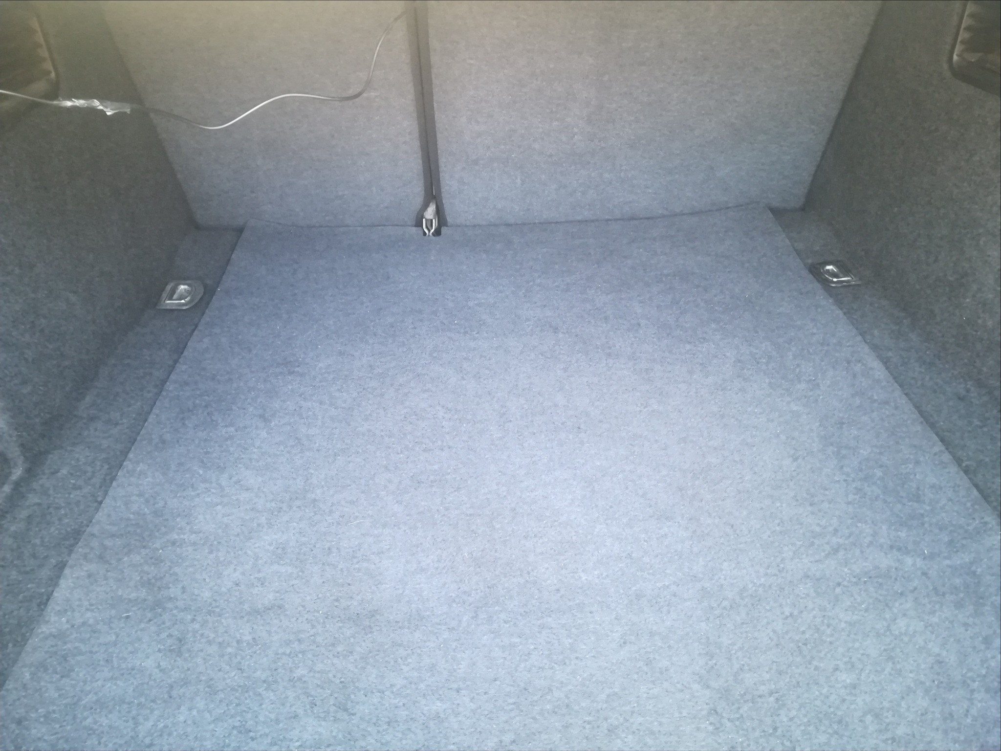 Szary, pusty bagażnik samochodu wyłożony filcem, z widocznymi metalowymi uchwytami mocującymi po bokach i centralnym zaczepem.