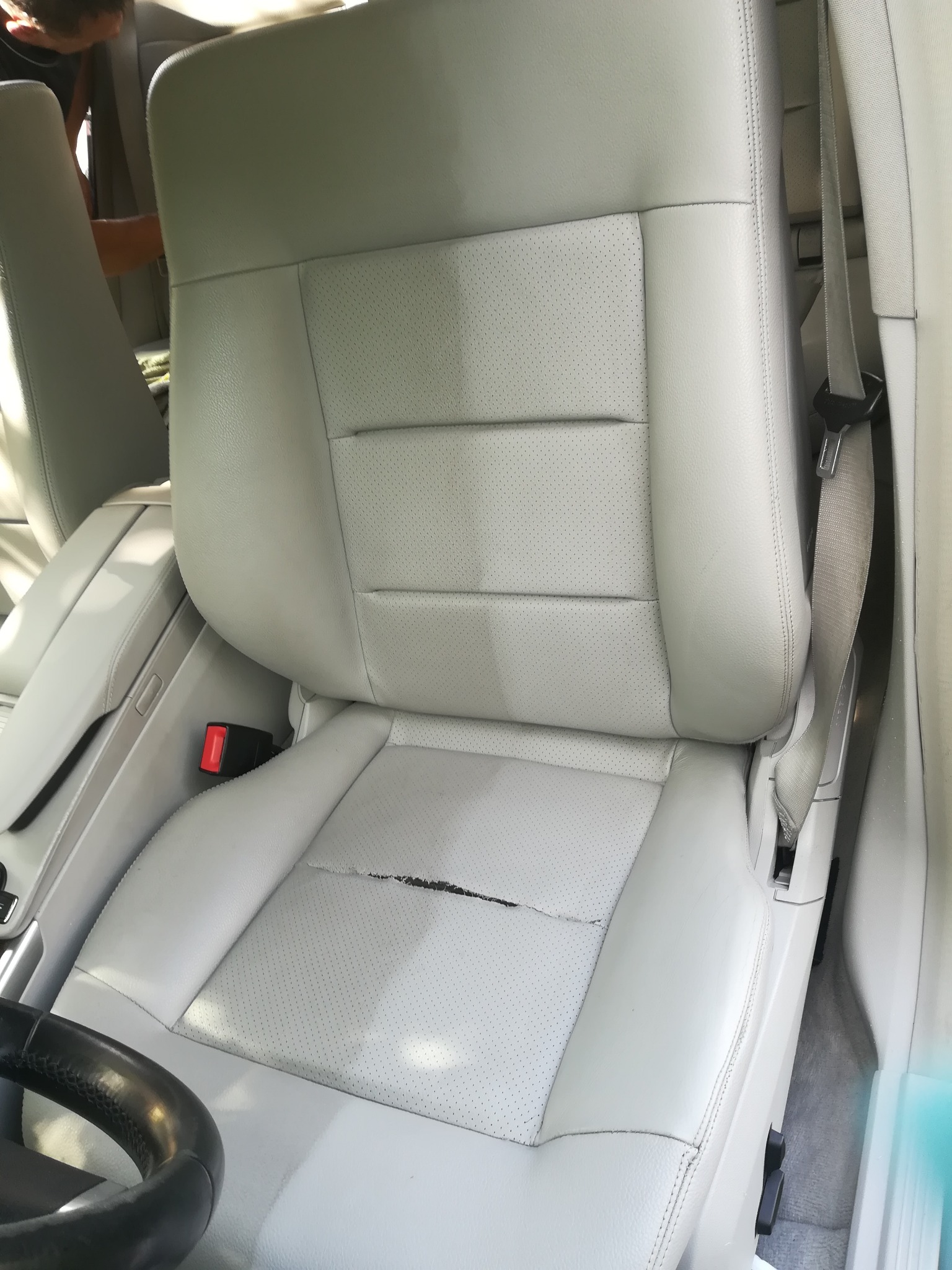 Jasnoszare skórzane siedzenie samochodowe z widocznym pęknięciem na siedzisku, widok z bliska, z fragmentem kierownicy i pasem bezpieczeństwa.