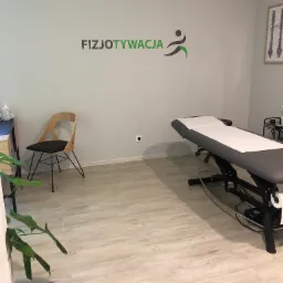Jasne wnętrze gabinetu z leżanką do masażu, krzesłem z drewnianym oparciem, plakatem z kręgosłupem i logo 'FIZJOTYWACJA' na ścianie.