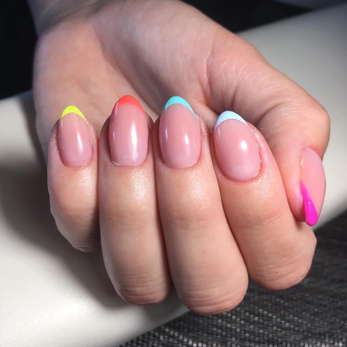 Dłoń z owalnymi paznokciami, każdy pomalowany na pastelowy kolor z neonową końcówką: żółtą, pomarańczową, błękitną, jasnoniebieską i różową.