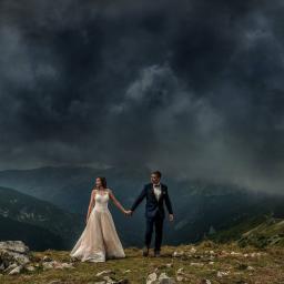 Foto Mono - Twój Ślub Marzeń - Para młoda, on w granatowym smokingu z muchą, ona w koronkowej sukni ecru, trzymają się za ręce na tle gór i burzowego nieba.