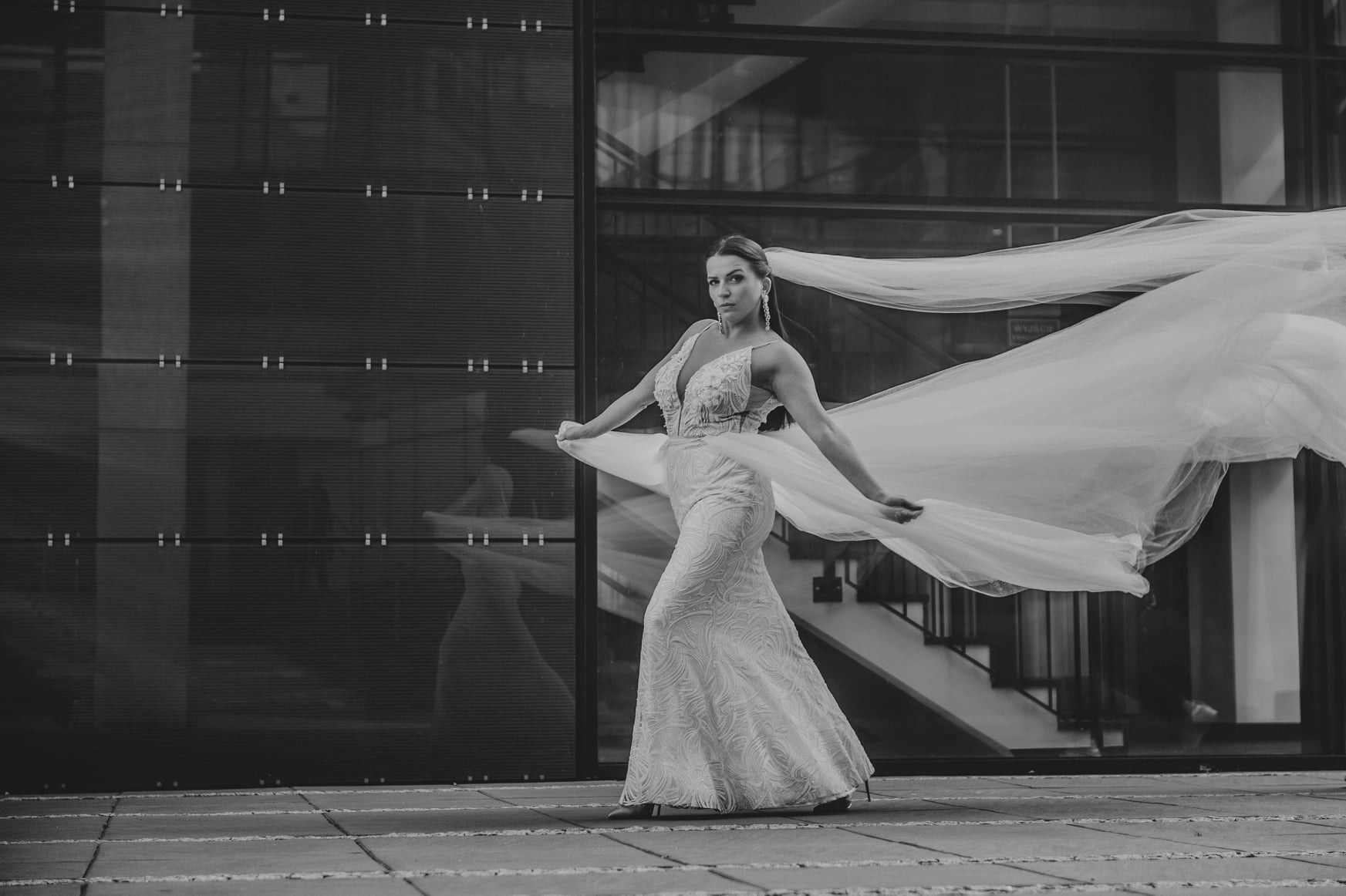 Elegancka kobieta w sukni ślubnej z długim welonem, pozująca na tle nowoczesnego budynku z szklaną fasadą, w monochromatycznej tonacji.
