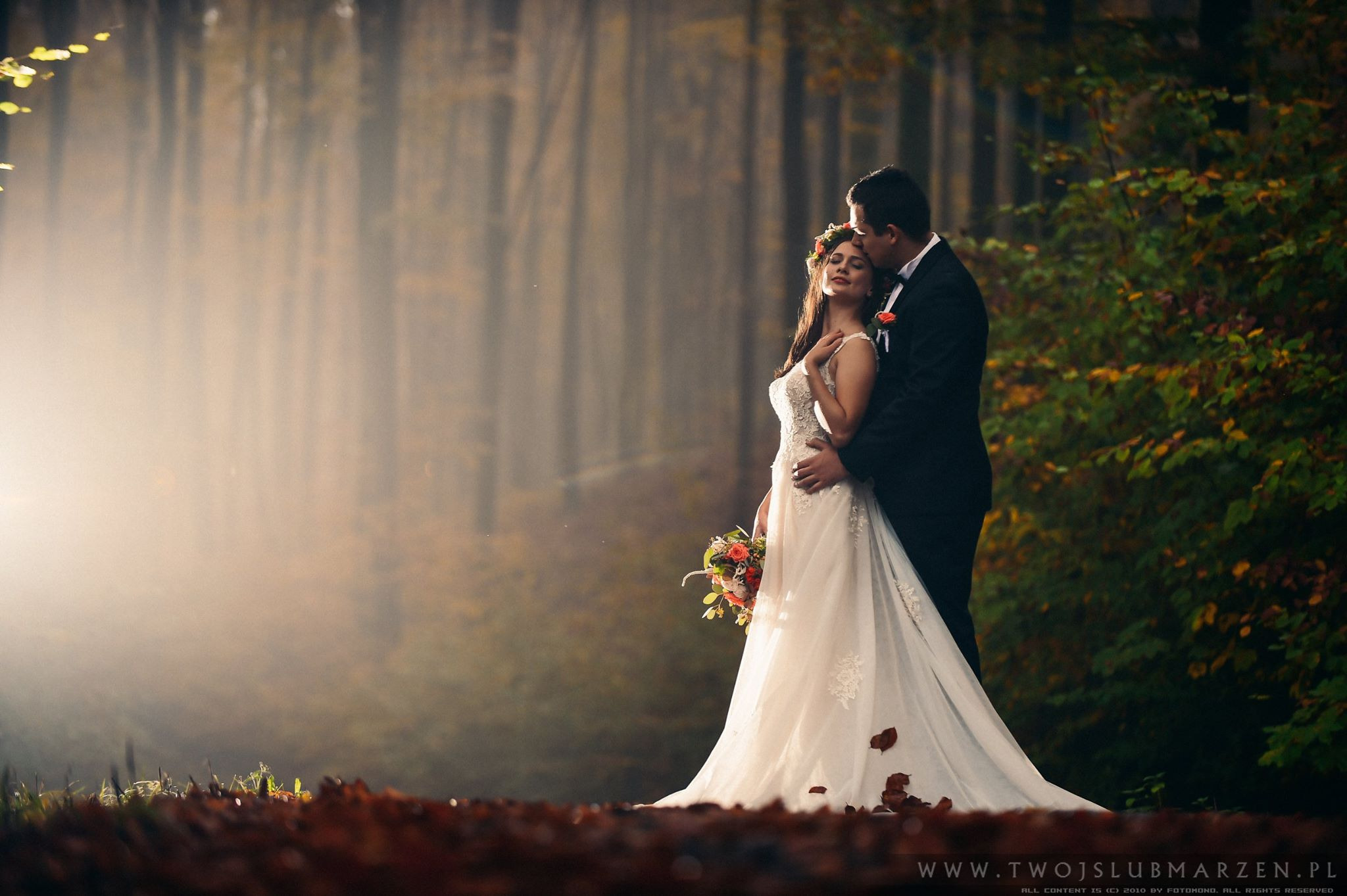 Para młoda w jesiennym lesie, mężczyzna w czarnym garniturze całuje kobietę w białej sukni ślubnej z kwiatowym wiankiem na głowie, promienie słońca przebijają przez mgłę.