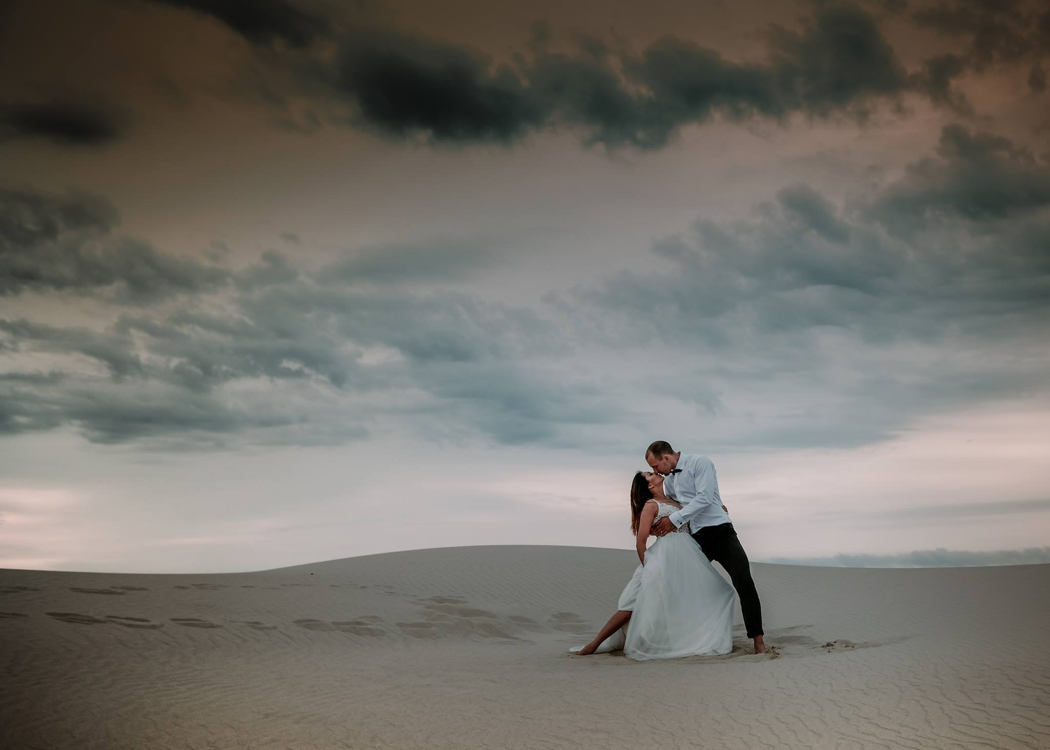 Para młoda w objęciach na piaszczystej wydmie pod pochmurnym niebem, on w białej koszuli i czarnych spodniach, ona w białej sukni ślubnej, widoczne ślady stóp na piasku.