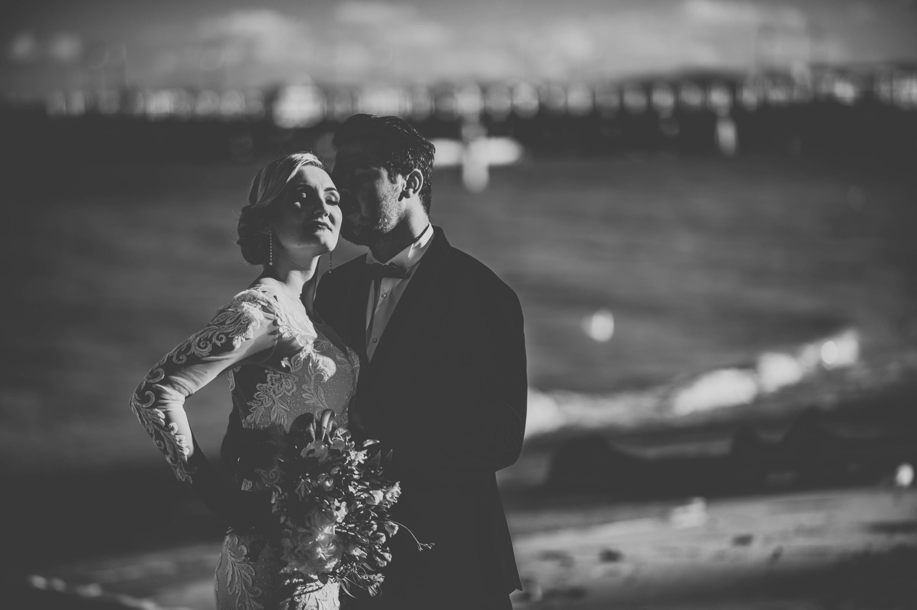 Czarno-białe zdjęcie nowożeńców: Panna Młoda w koronkowej sukni z bukietem, Pan Młody w smokingu, delikatnie całujący ją w policzek na tle rozmytego krajobrazu morskiego.