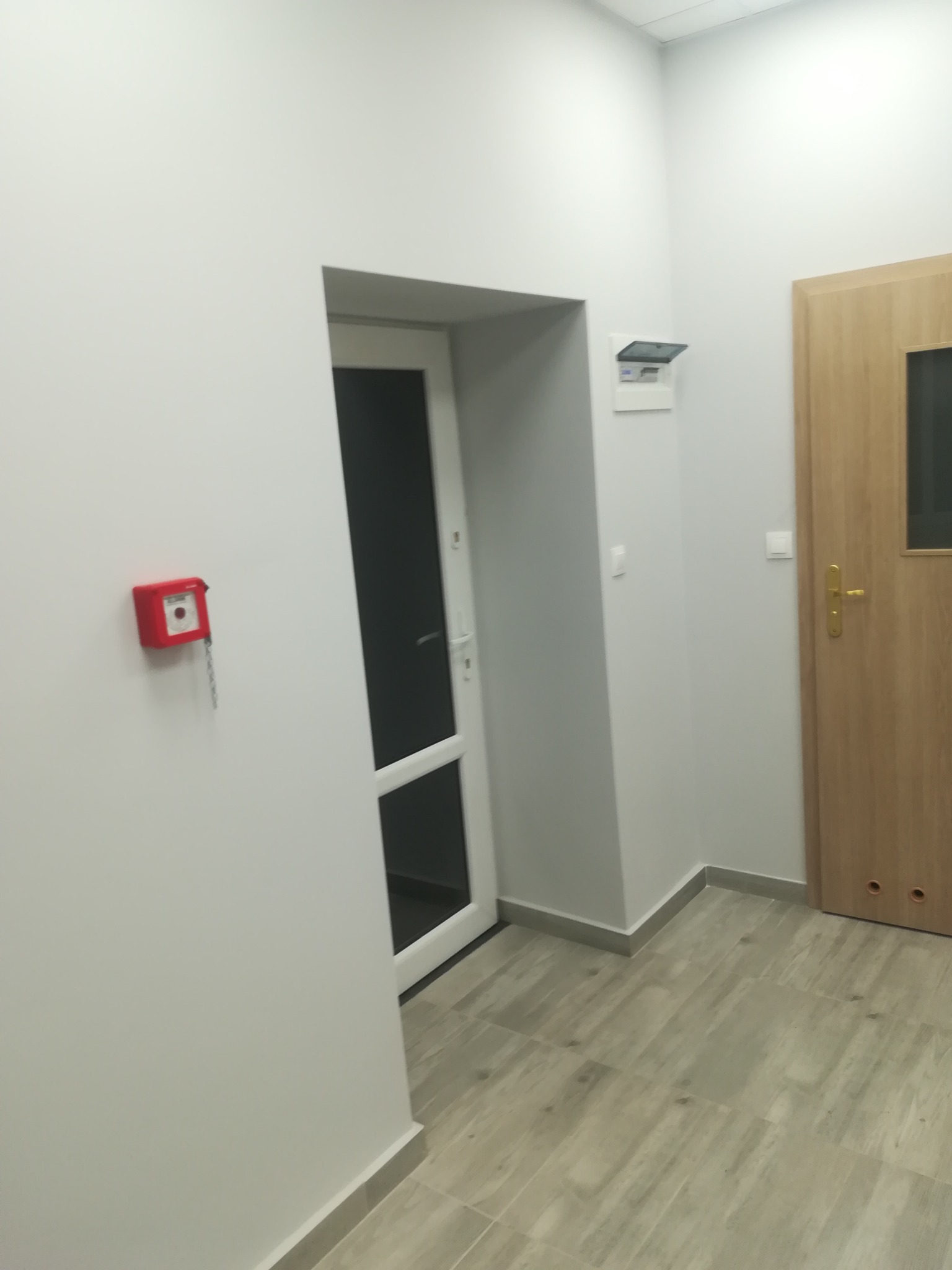 Wnętrze biurowe: Jasne ściany, drewnopodobne płytki podłogowe, białe drzwi z przeszkleniem, brązowe drzwi z klamką, czerwony alarm przeciwpożarowy na ścianie i skrzynka elektryczna z otwartą klapką.