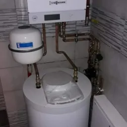 Nowy, biały, wiszący piec gazowy Viessmann z miedzianą instalacją, zbiornikiem wyrównawczym i zasobnikiem ciepłej wody, zabezpieczonym folią ochronną, na tle płytek ceramicznych.