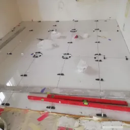 Białe płytki podłogowe w trakcie układania, z krzyżykami dystansowymi i poziomnicami, widoczne ślady kleju i zaprawy.