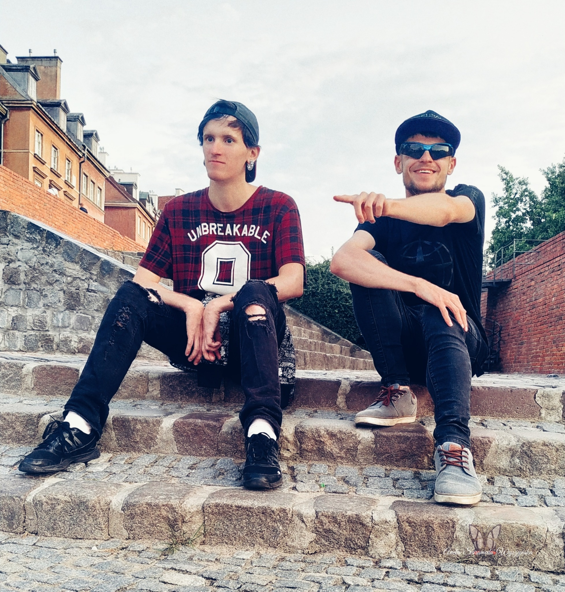 Dwaj młodzi mężczyźni siedzą na kamiennych schodach w miejskim otoczeniu, jeden w czapce i okularach słonecznych wskazuje palcem w bok, drugi w czapce z kolczykiem patrzy w dal.