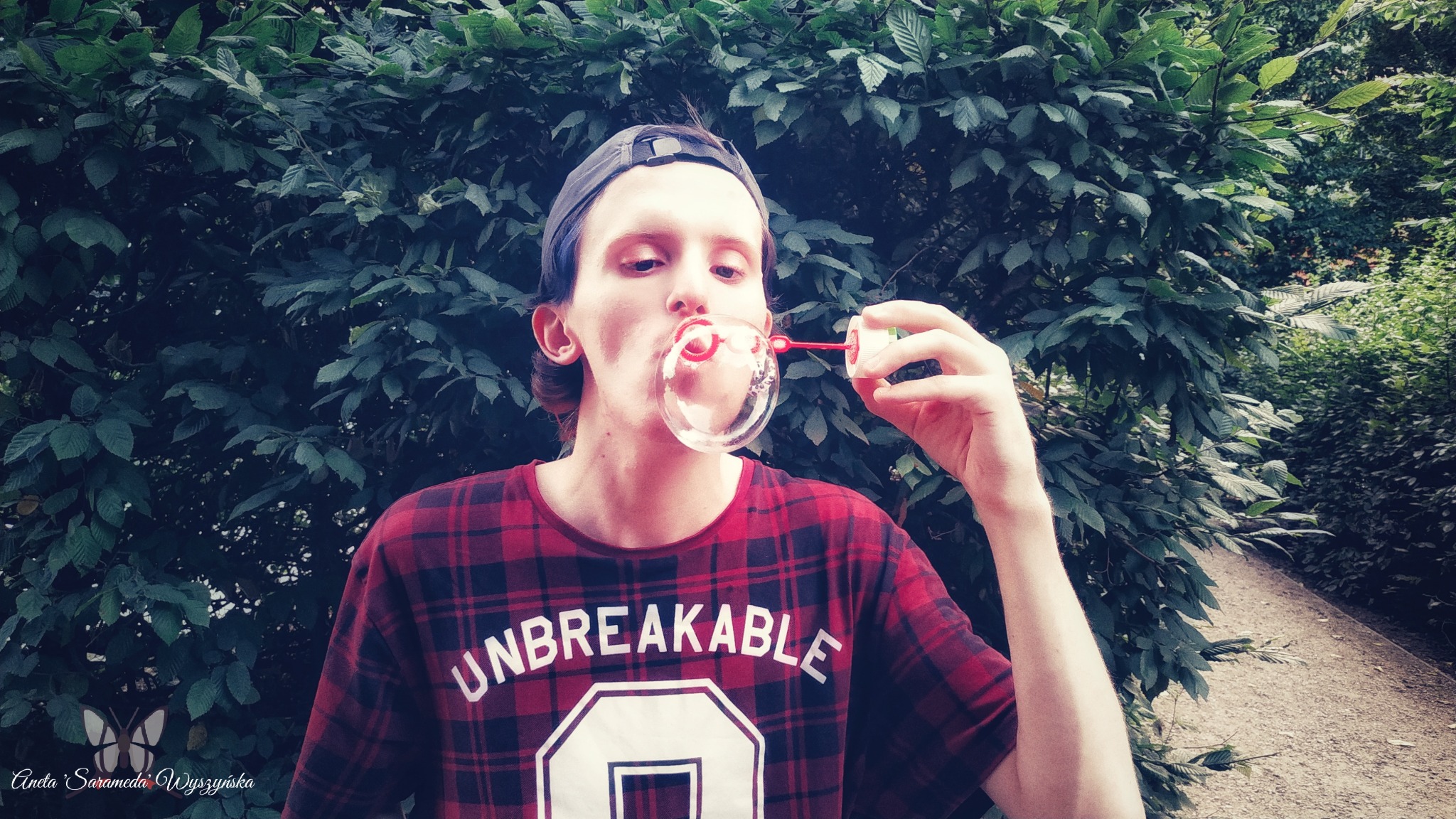 Młody mężczyzna w czapce i koszuli w kratę dmucha bańki mydlane na tle gęstych zielonych liści.