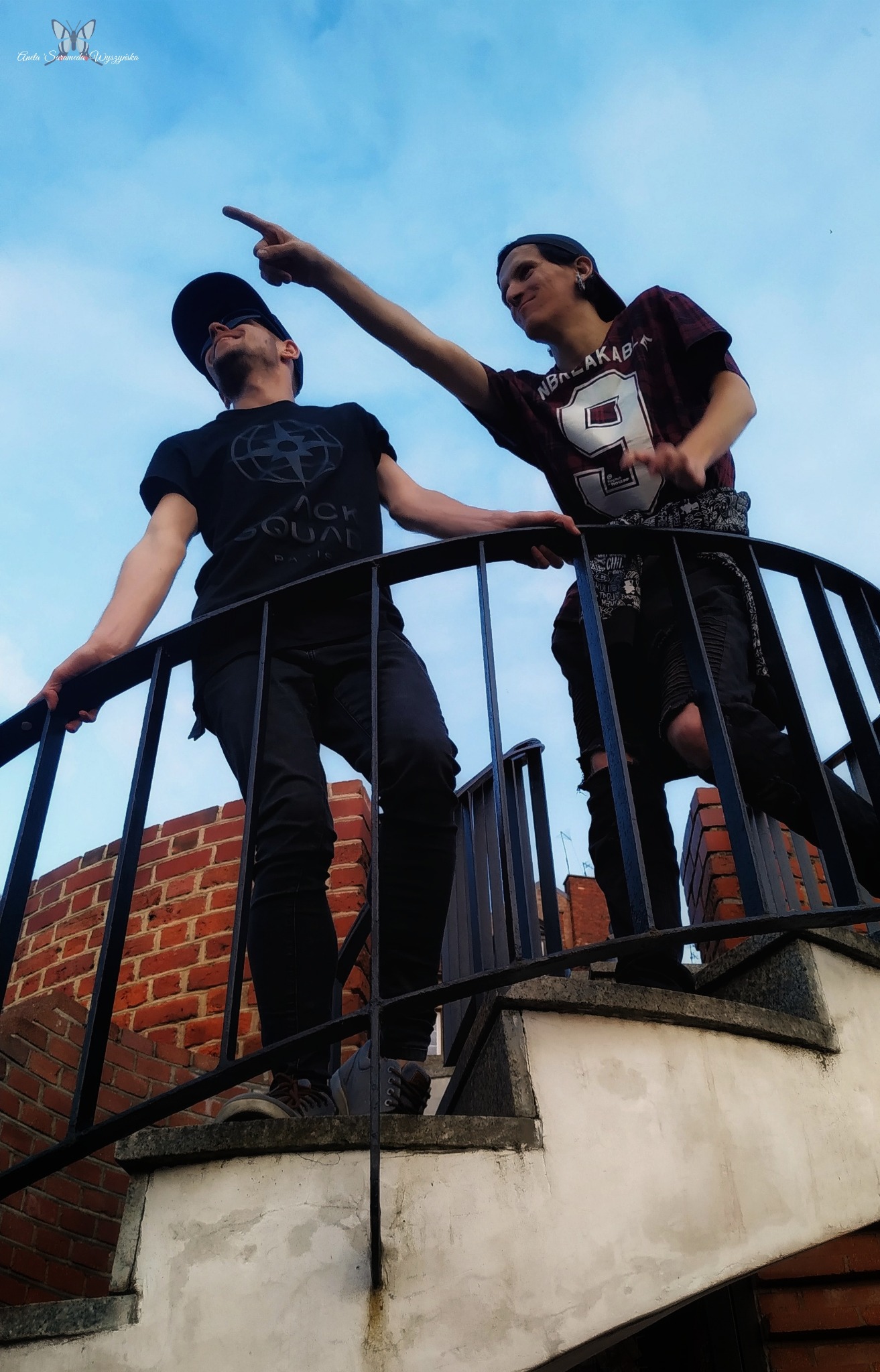 Dwóch młodych mężczyzn w casualowych strojach pozuje na krętych schodach na tle błękitnego nieba, jeden wskazuje palcem w górę.