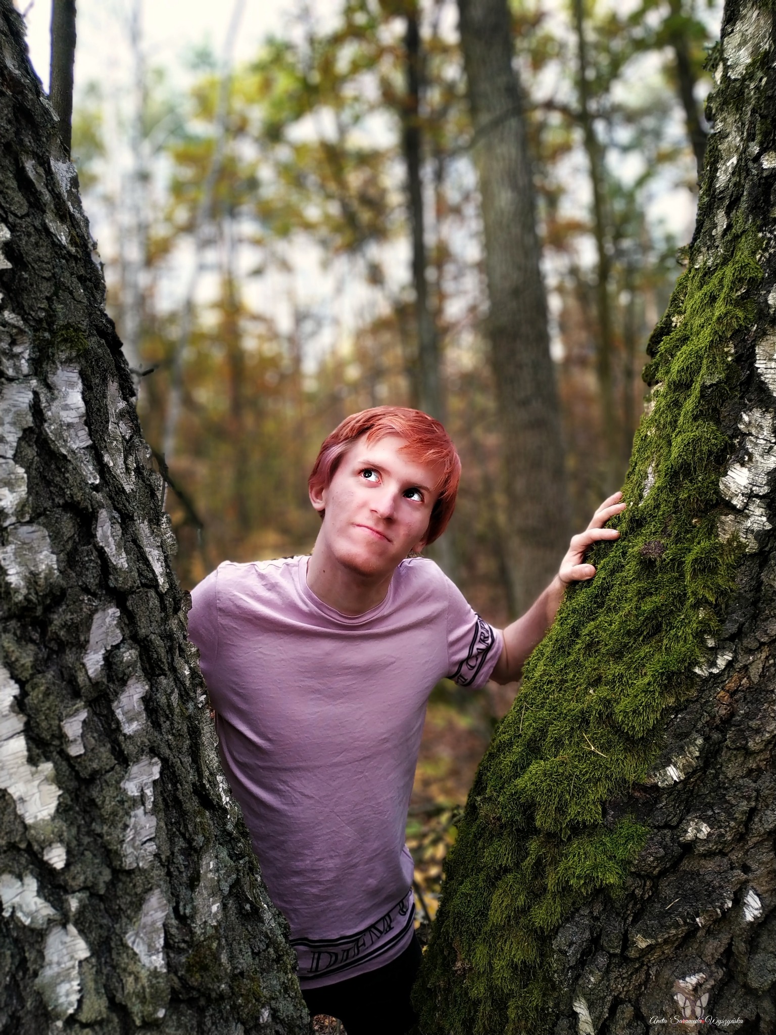 Młody mężczyzna z rudymi włosami w liliowej koszulce opiera się o dwa drzewa porośnięte mchem, spoglądając w górę w lesie.