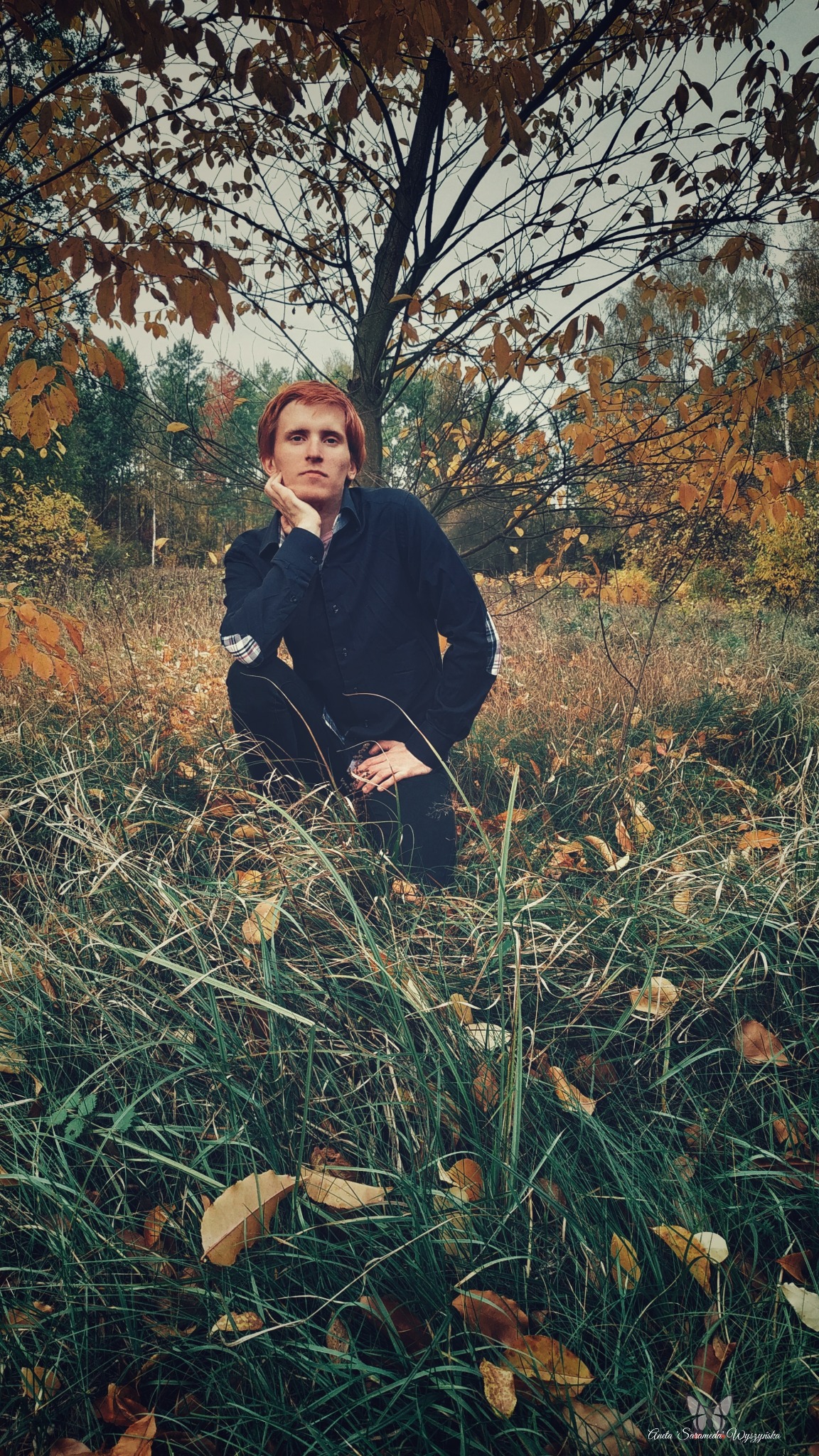 Mężczyzna o rudych włosach kuca w wysokiej trawie na tle jesiennego lasu, opierając brodę na dłoni i spoglądając w obiektyw.