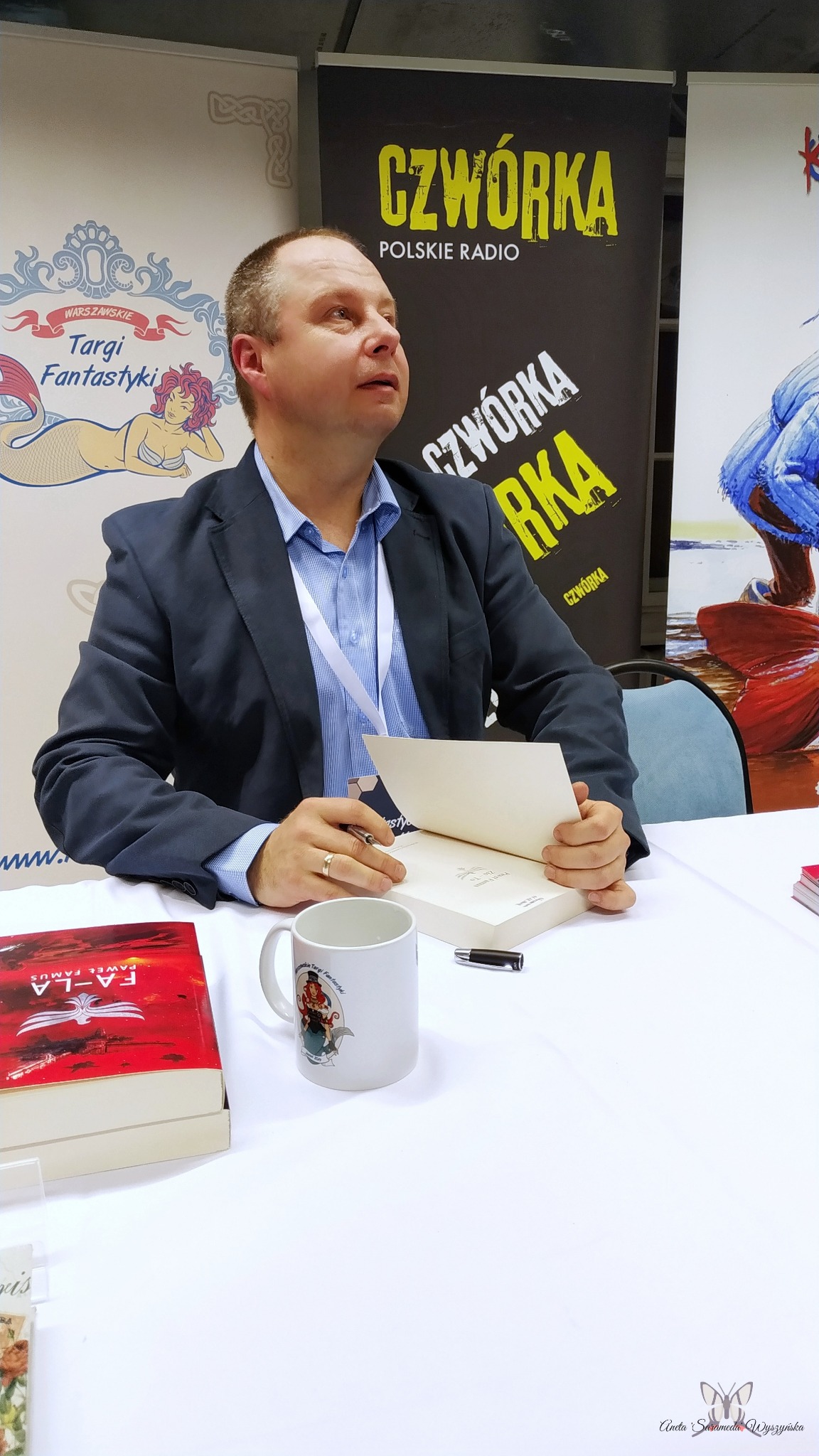 Mężczyzna w garniturze siedzi za stołem, podpisując książkę na tle banerów reklamowych, w pobliżu kubek z logo Targów Fantastyki.