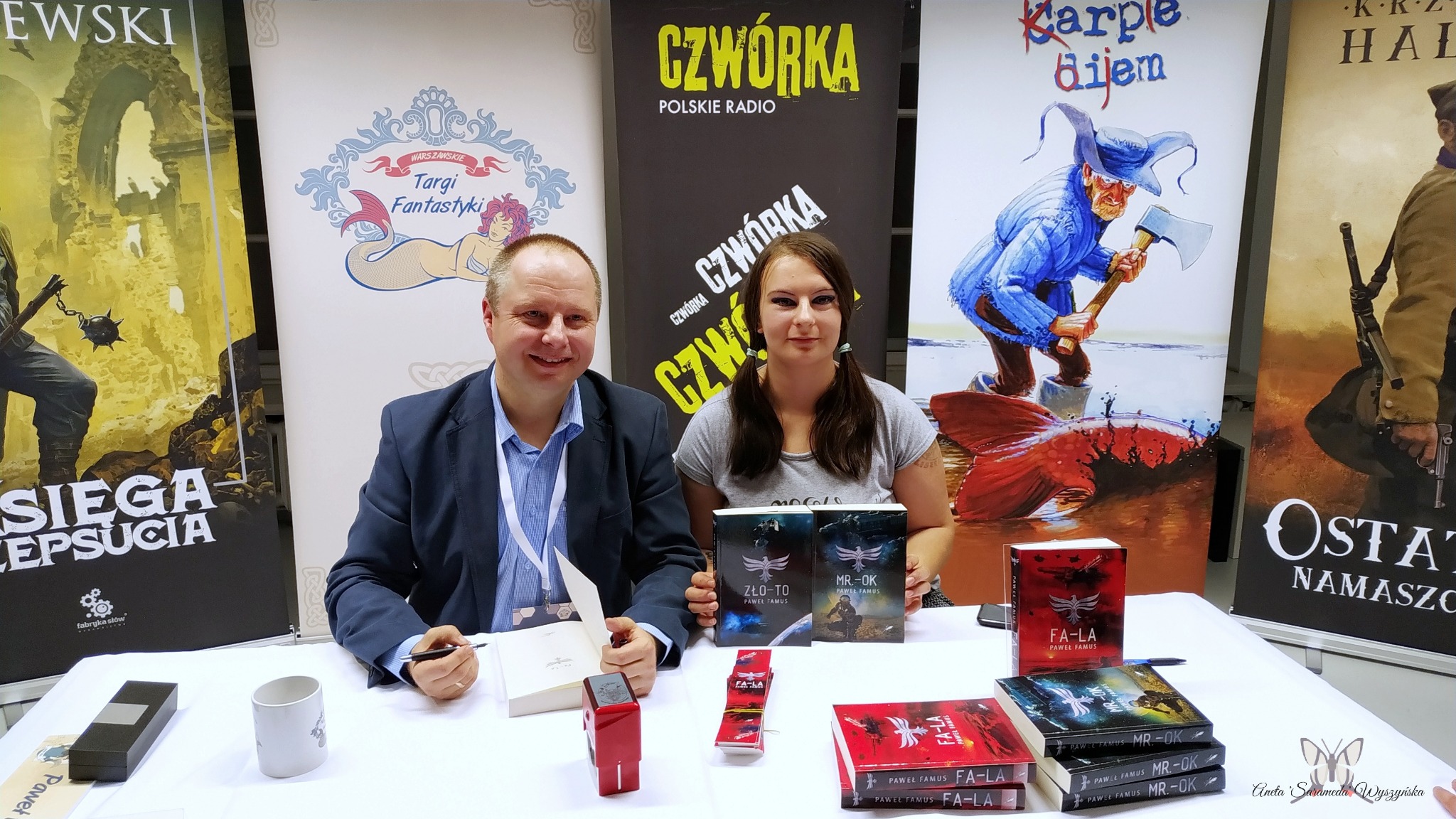 Autorzy książek fantastycznych, mężczyzna w marynarce podpisuje egzemplarz, obok kobieta prezentuje okładki powieści na tle banerów reklamowych targów książki.