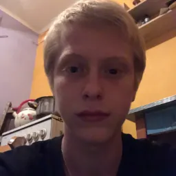 Portret młodej osoby z blond włosami na tle kuchni z kuchenką i półkami.