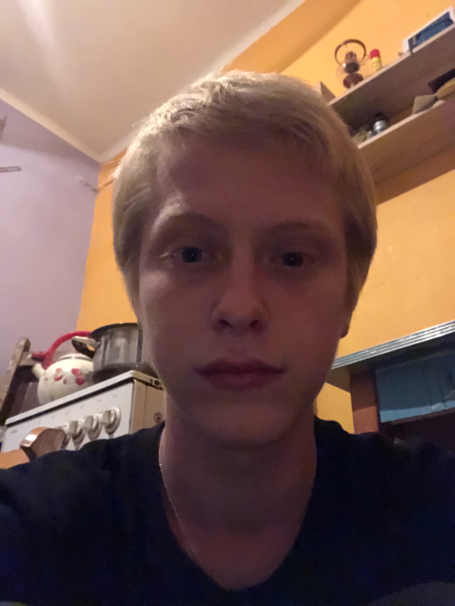 Portret młodej osoby z blond włosami na tle kuchni z kuchenką i półkami.