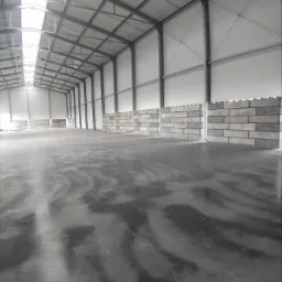 Wnętrze hali z betonową posadzką i częściowo wymurowanymi ścianami z bloczków betonowych, widoczne stalowe elementy konstrukcyjne dachu i świetliki.