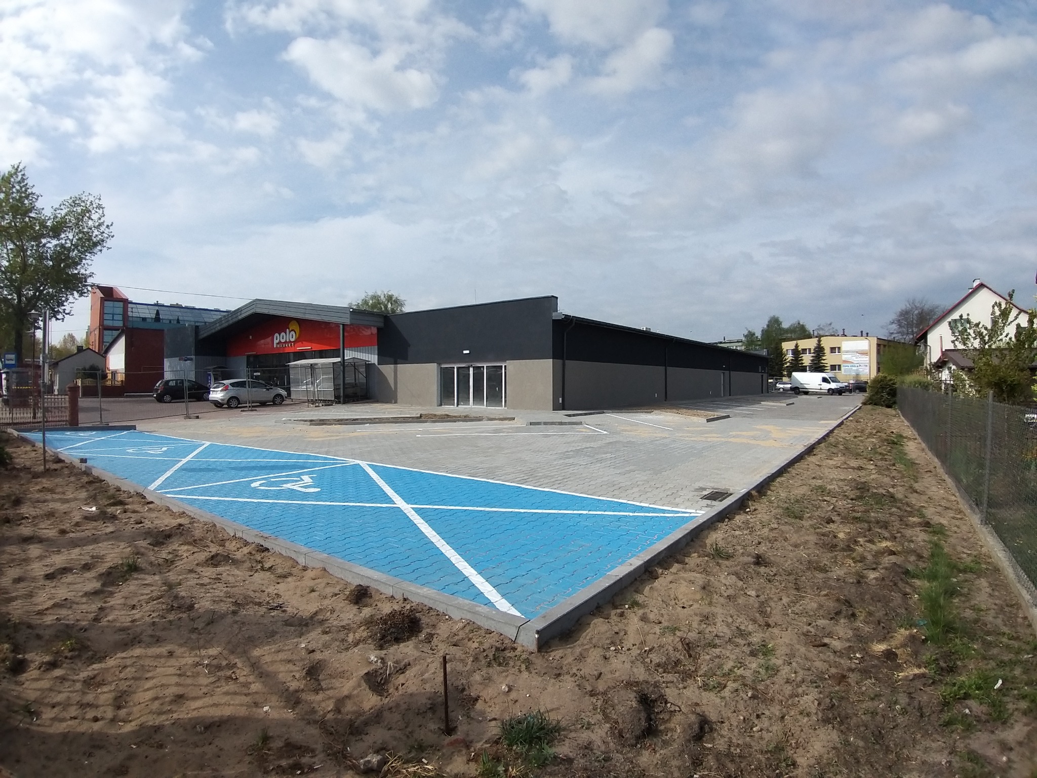 Nowo wybrukowany parking z niebieską kostką dla osób niepełnosprawnych przed budynkiem sklepu Polo Market, widoczne samochody i fragment ogrodzenia.