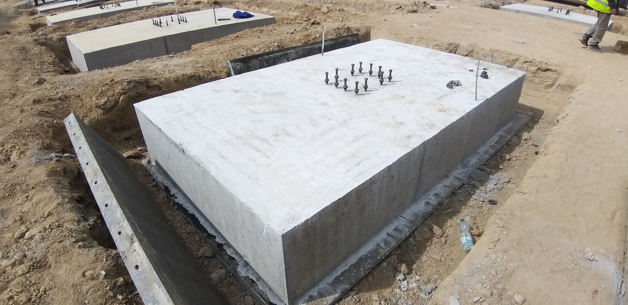 Betonowe fundamenty w wykopie, z wystającymi śrubami kotwiącymi, widoczna butelka wody obok jednego z fundamentów.