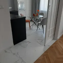 Nowoczesna kuchnia z białymi szafkami, czarnym blatem, szklanym stołem i krzesłami w odcieniach szarości, widok z perspektywy podłogi z marmurowymi płytkami przechodzącej w drewniany parkiet...