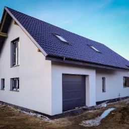 Nowoczesny, dwukondygnacyjny dom z ciemną dach&oacute;wką i garażem w bryle, w trakcie wykończenia elewacji zewnętrznej, widoczne fragmenty ocieplenia i przygotowania terenu.