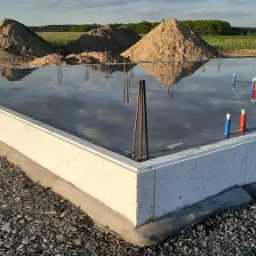 Zalana płyta fundamentowa z wystającymi prętami zbrojeniowymi i rurami instalacyjnymi, na tle pryzm piasku i zieleni.
