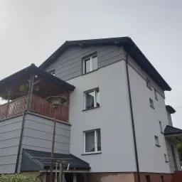 Dwupiętrowy dom z balkonem, elewacja w odcieniach szarości, widoczne świeżo pomalowane ściany i elementy dekoracyjne.