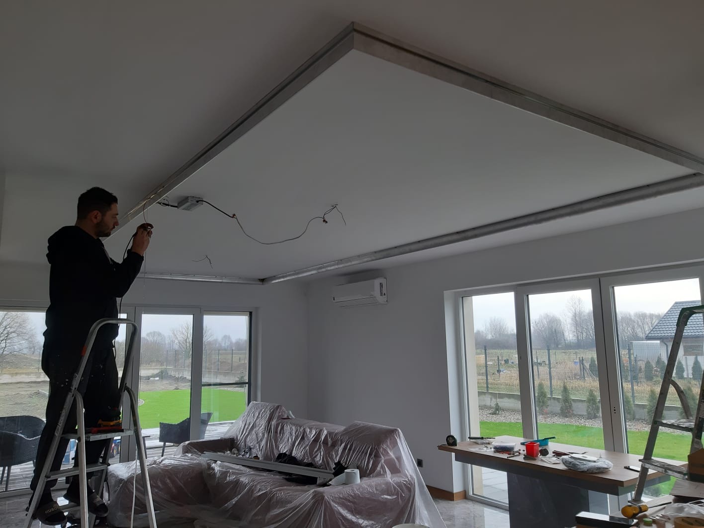 Elektryk na drabinie łączy przewody w suficie podwieszanym z płyt karton gips w jasnym salonie z dużymi oknami i meblami zabezpieczonymi folią.