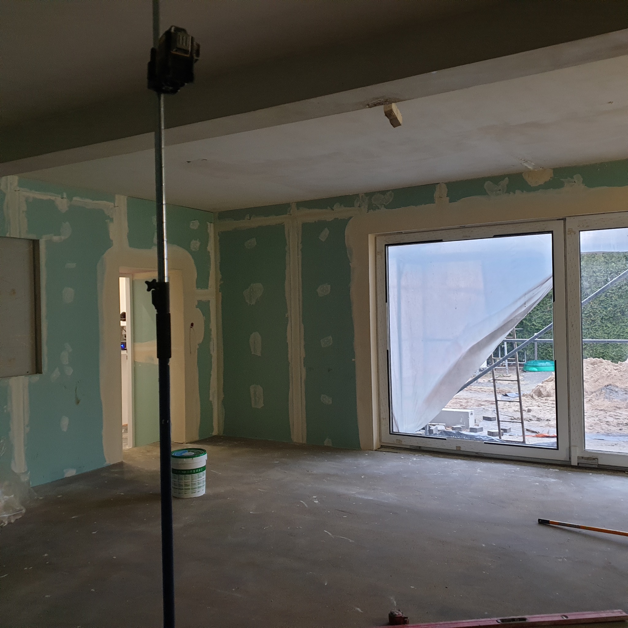 Wnętrze w trakcie remontu: ściany pokryte płytami kartonowo-gipsowymi, częściowo zaszpachlowane, okno zasłonięte folią ochronną, widoczny laser krzyżowy na statywie i wiadro z farbą.