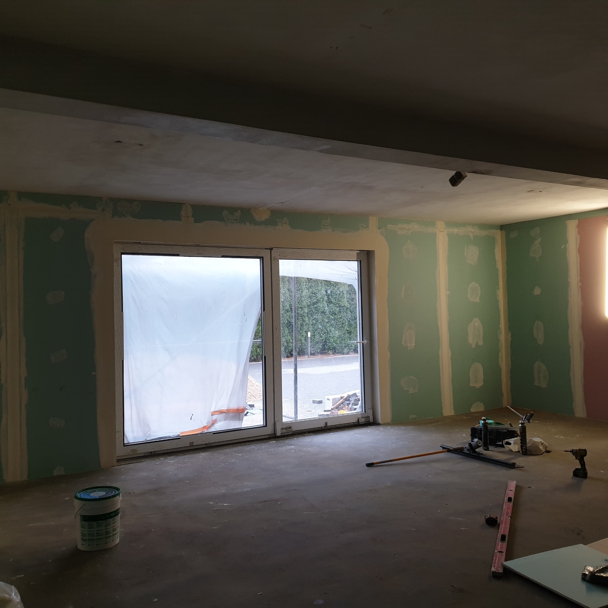 Wnętrze w trakcie prac wykończeniowych: ściany z płyt kartonowo-gipsowych, częściowo wyszpachlowane, okno zabezpieczone folią, widoczne narzędzia i wiadro z farbą na podłodze.