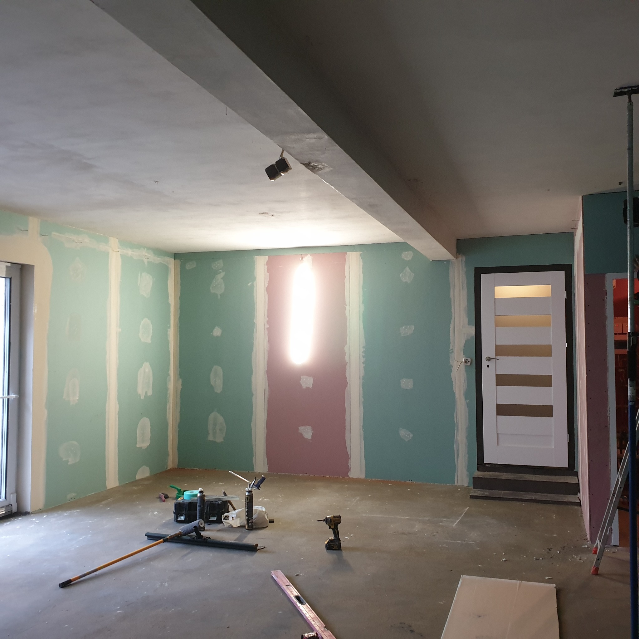Wnętrze w trakcie prac wykończeniowych: ściany z płyt kartonowo-gipsowych, częściowo wyszpachlowane, widoczne narzędzia i poziomica na podłodze, przygotowane do dalszych prac.