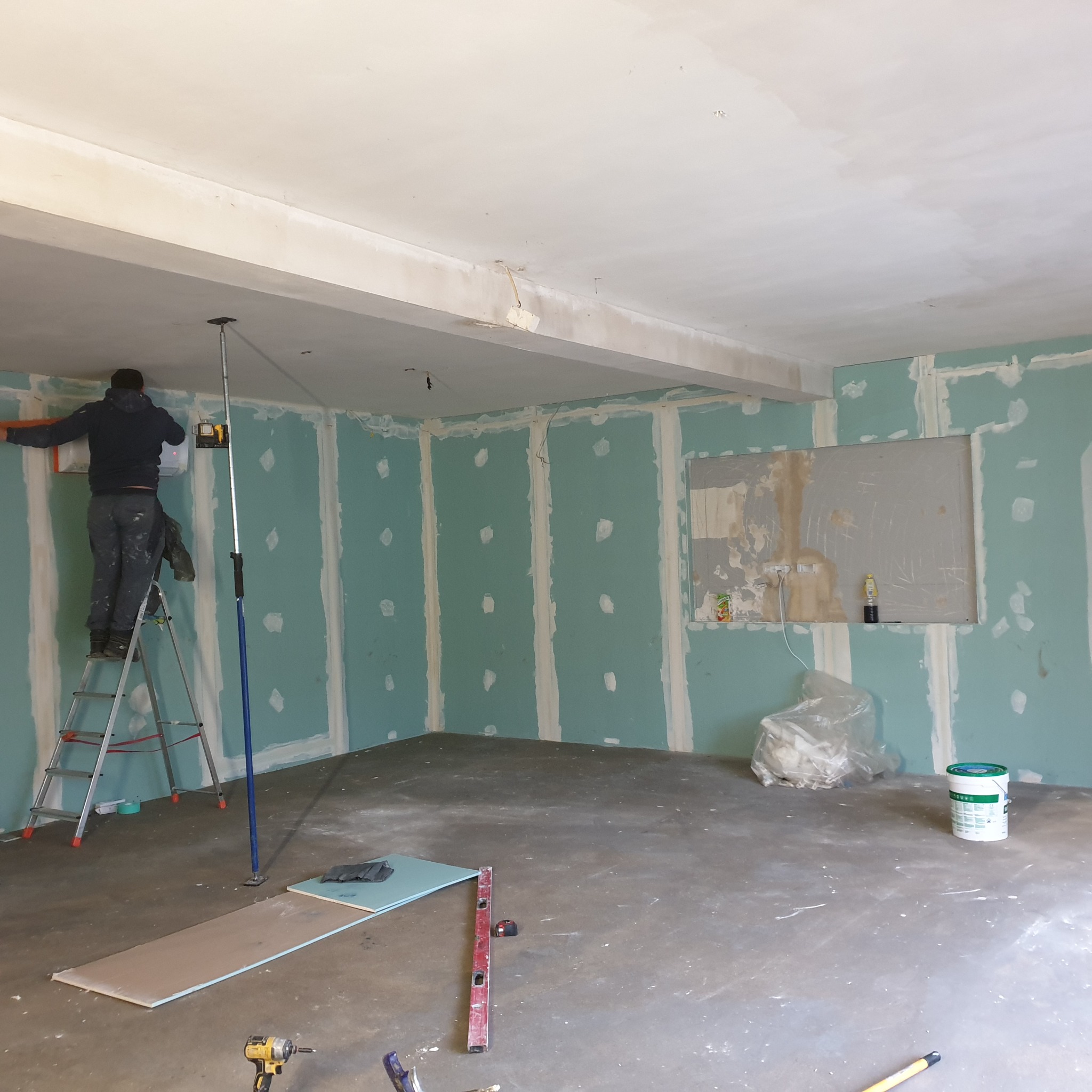 Pracownik na drabinie montuje płyty kartonowo-gipsowe na ścianie, widoczny laser krzyżowy, narzędzia i materiały budowlane na podłodze w surowym pomieszczeniu.