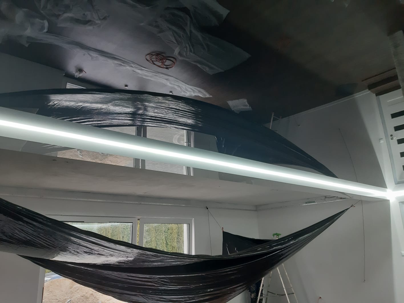 Przygotowanie pomieszczenia do prac wykończeniowych: sufit i ściany zabezpieczone folią, oświetlenie listwowe LED zamontowane, widoczna drabina i okno z widokiem na zieleń.