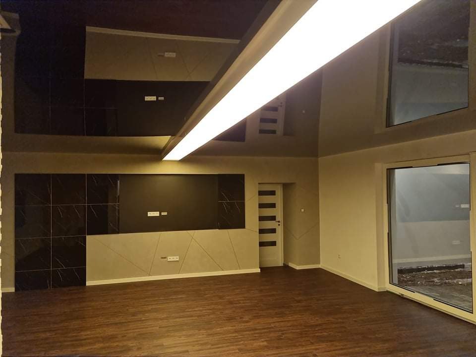 Nowoczesne wnętrze po remoncie: parkiet, ściany w odcieniach beżu i szarości, dekoracyjny panel ścienny z czarnych płytek imitujących marmur, sufit z połyskliwą, ciemną powierzchnią odbijającą...