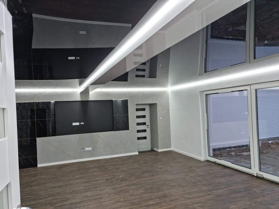 Nowoczesne wnętrze salonu z podświetlanymi panelami ściennymi w odcieniach szarości i czerni, drewnopodobną podłogą i dużymi oknami tarasowymi.