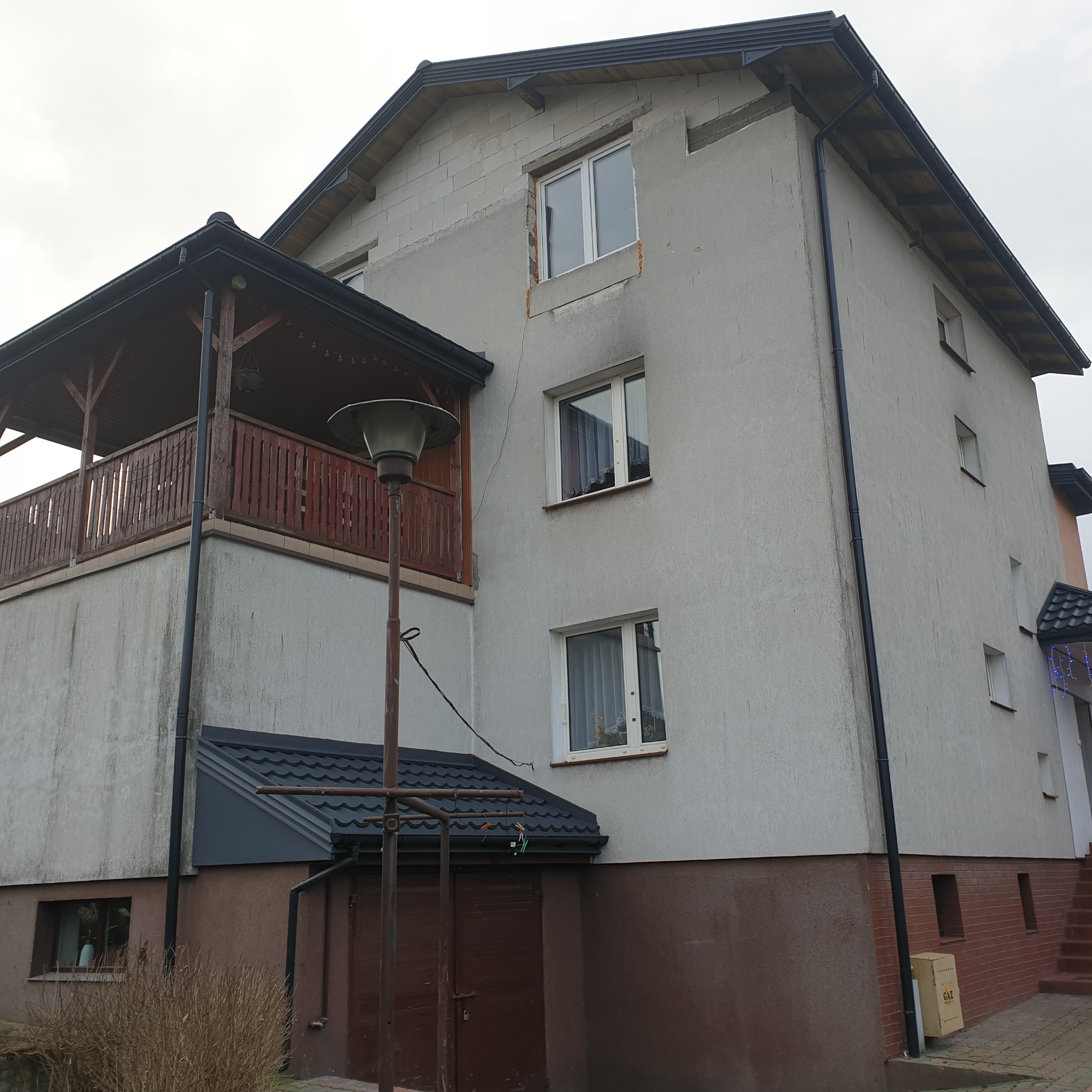 Dwukondygnacyjny dom z częściowo otynkowaną elewacją, widoczne okna z białymi ramami, drewniany balkon z balustradą, oraz fragment dachu pokrytego blachodachówką.