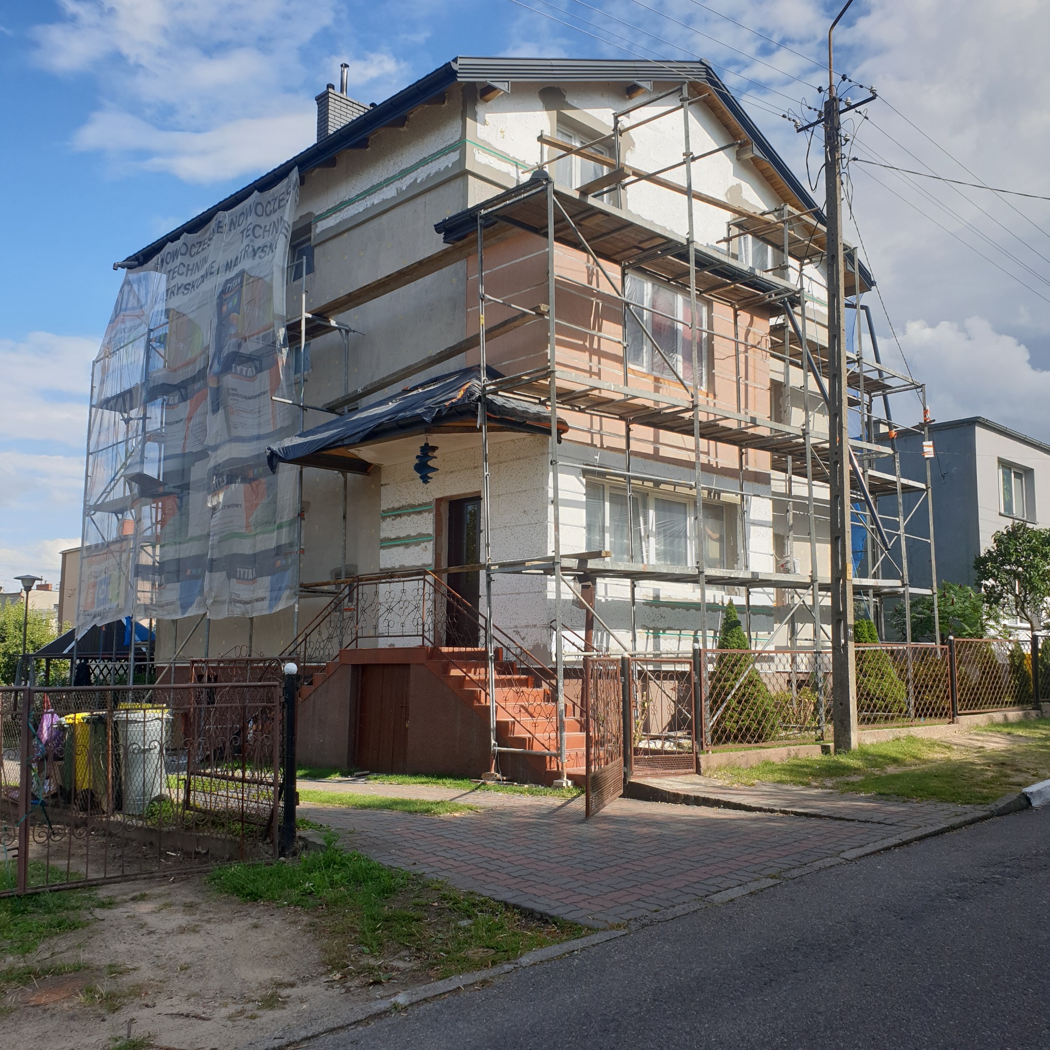 Dwupiętrowy dom w trakcie prac elewacyjnych, osłonięty siatką z reklamą materiałów budowlanych, z widocznym rusztowaniem, częściowo ocieplony, z elementami elewacji w kolorze białym i łososiowym.