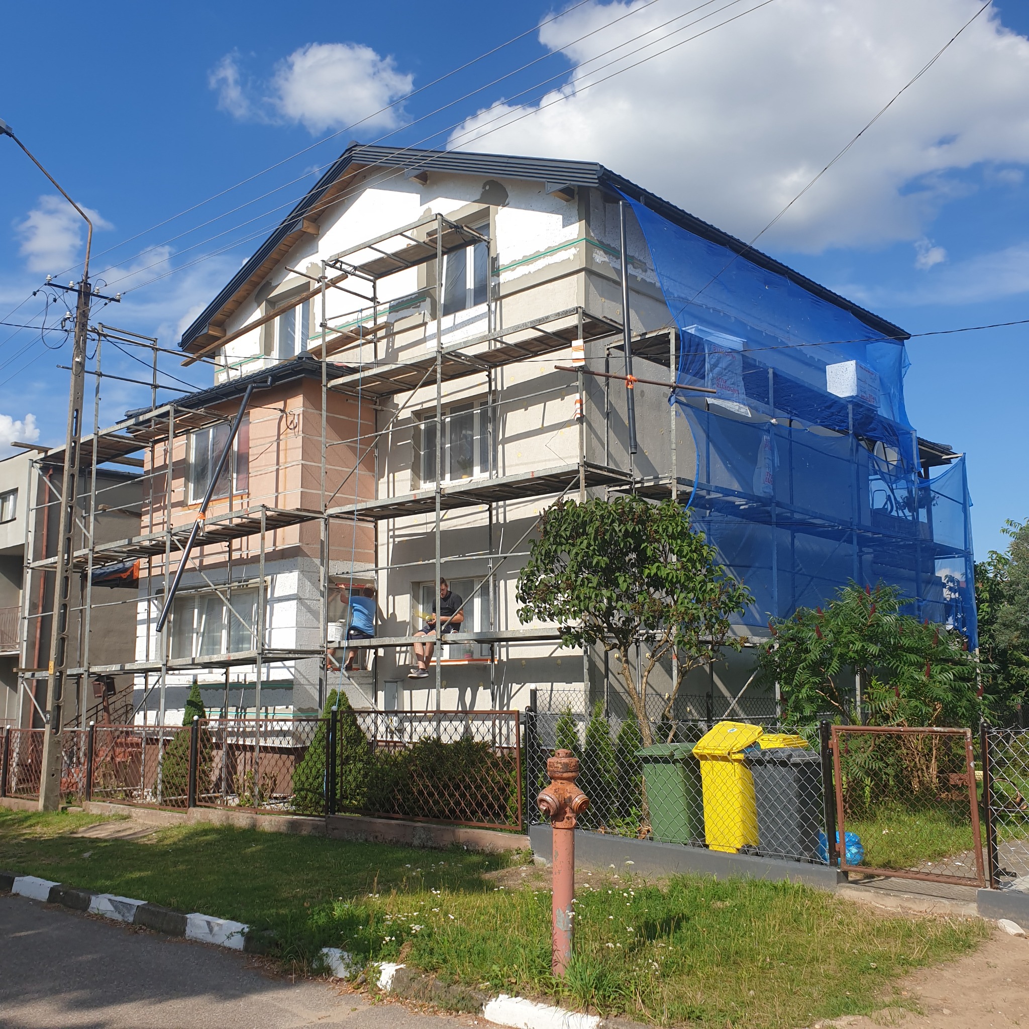 Dwupiętrowy dom w trakcie ocieplania, otoczony rusztowaniami i zabezpieczony niebieską siatką ochronną, widoczni pracownicy na rusztowaniu, hydrant i kosze na śmieci na pierwszym planie.