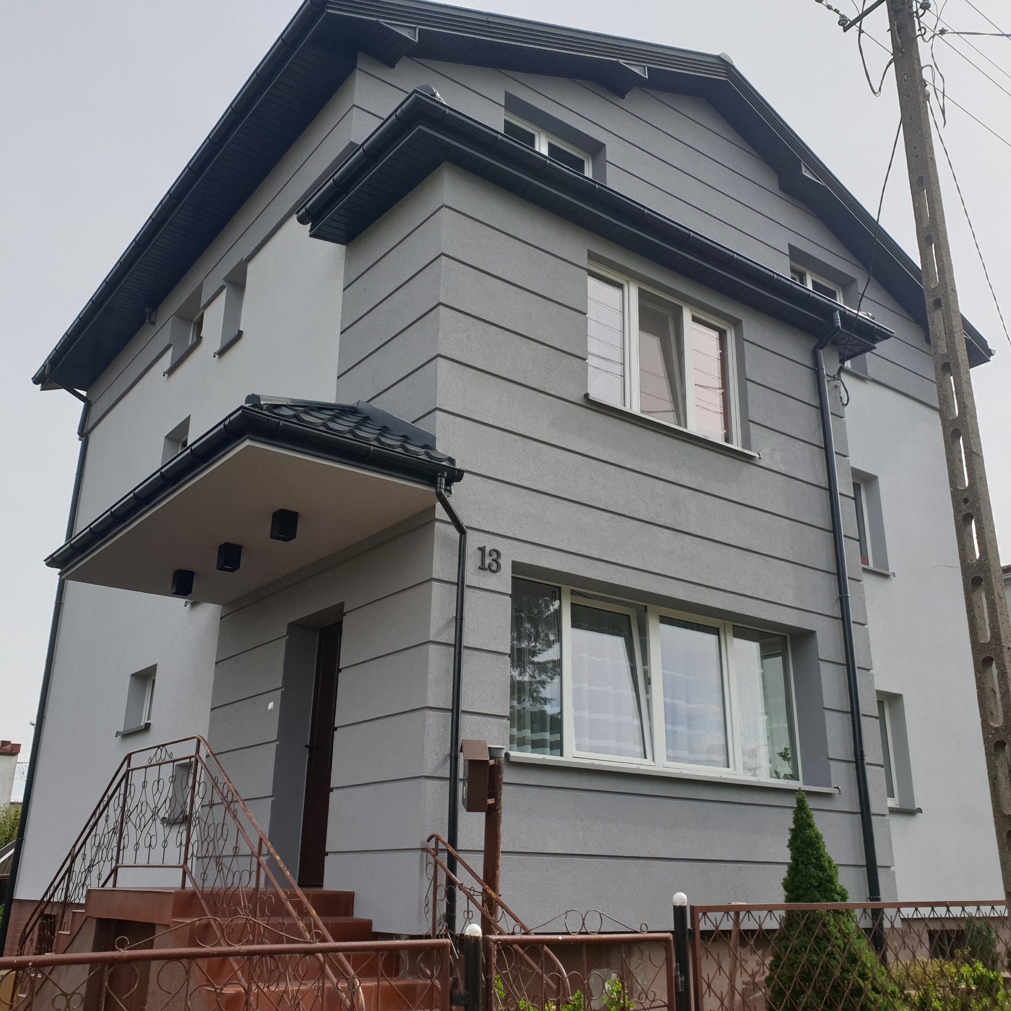 Dwukondygnacyjny dom jednorodzinny z elewacją w odcieniach szarości, z numerem '13' nad drzwiami wejściowymi i metalowymi schodami z dekoracyjną balustradą.