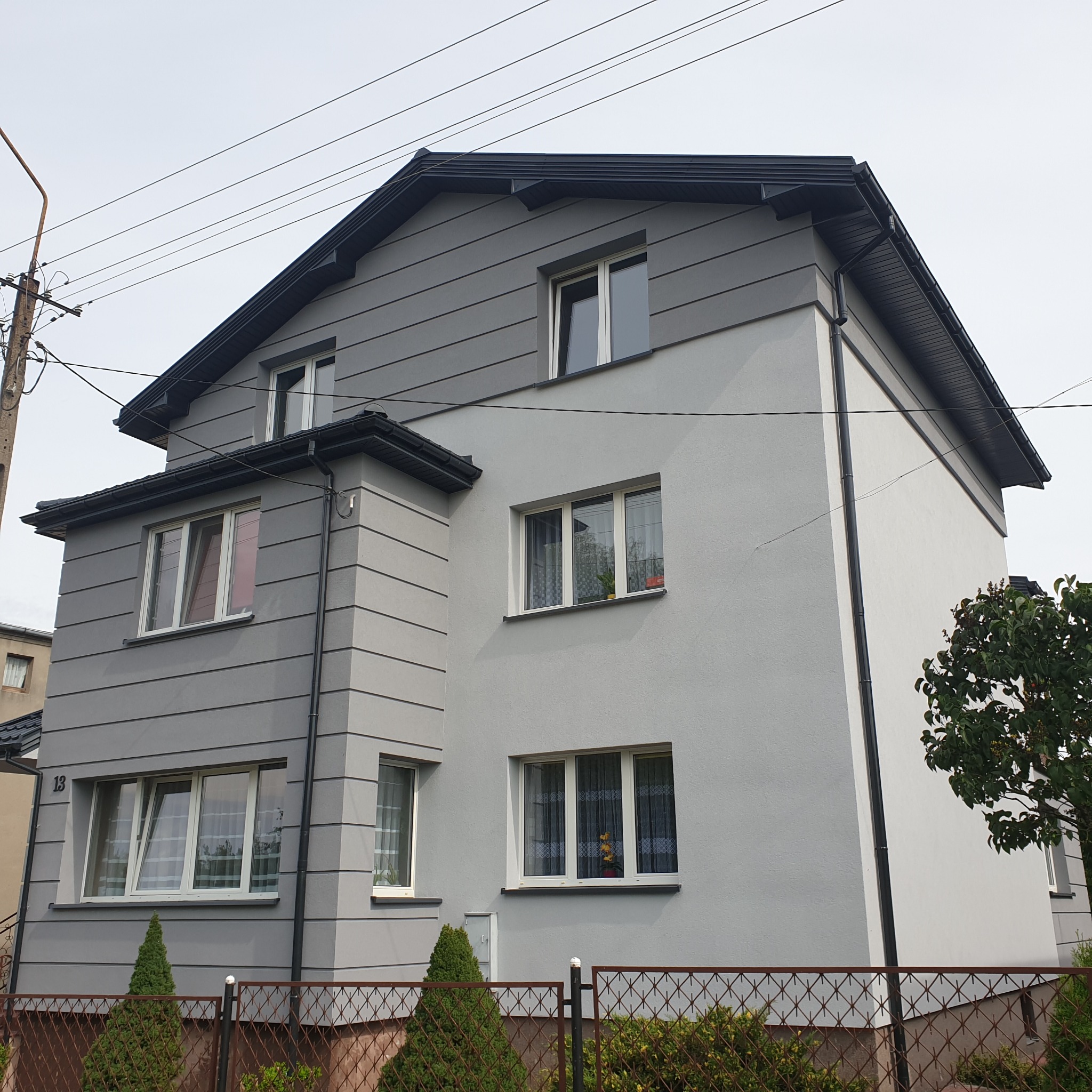 Dwupiętrowy dom jednorodzinny po remoncie elewacji, z szarą, poziomą okładziną imitującą panele na górnej części i gładkim, jasnoszarym tynkiem na dolnej, widoczny zza brązowego ogrodzenia z siatki.