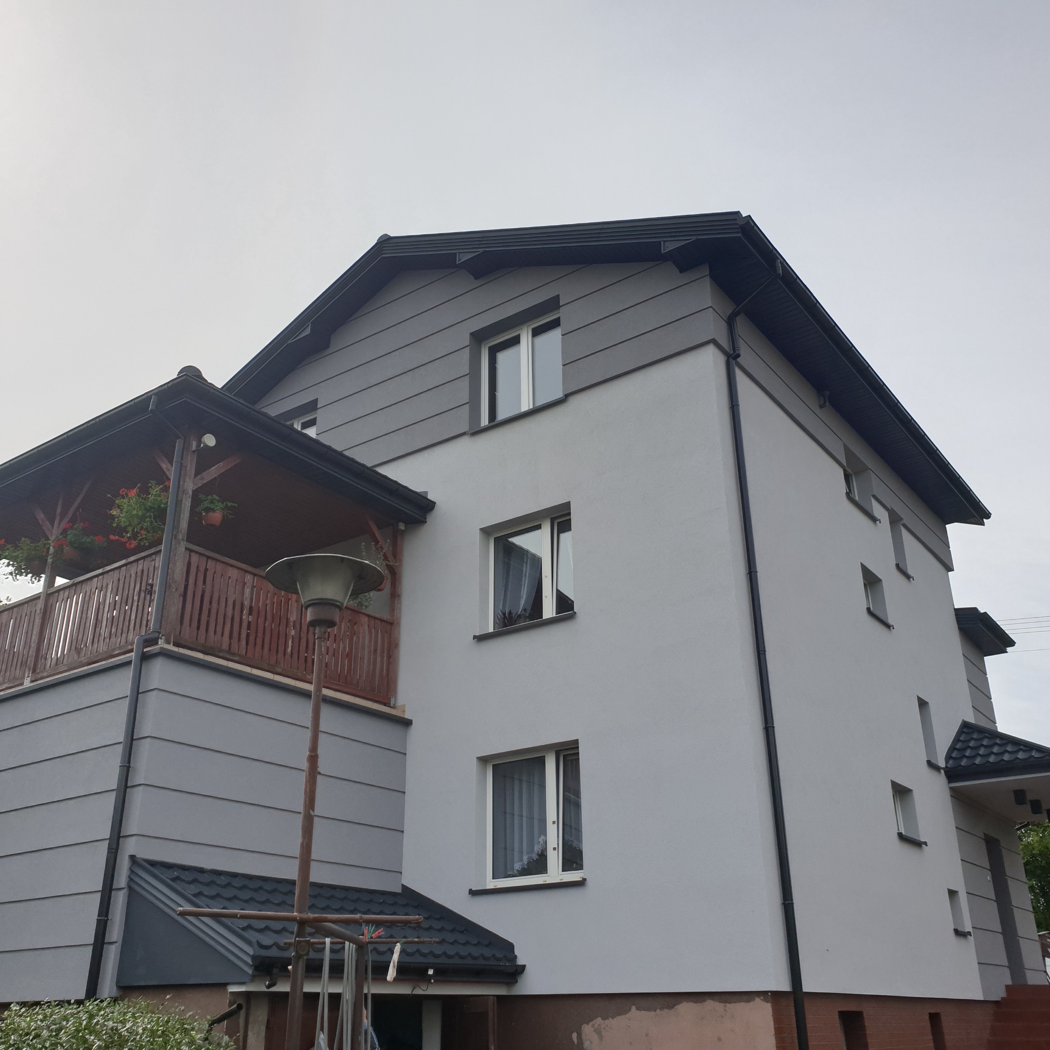 Dwupiętrowy dom z balkonem, elewacja w odcieniach szarości, widoczne świeżo pomalowane ściany i elementy dekoracyjne.