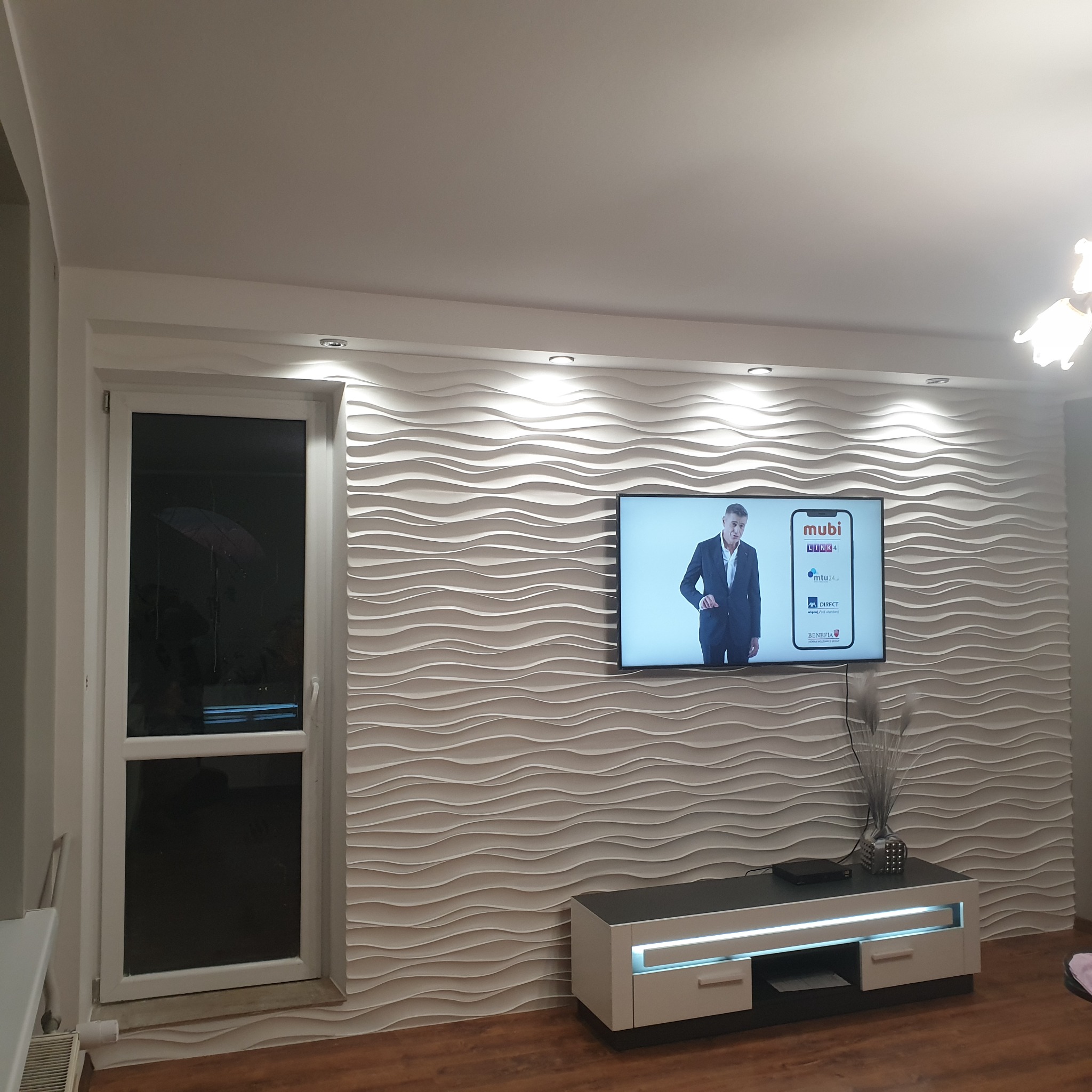 Salon z dekoracyjną ścianą 3D w odcieniach bieli, podświetloną punktowymi lampami sufitowymi, z telewizorem i nowoczesną szafką RTV z podświetleniem LED.