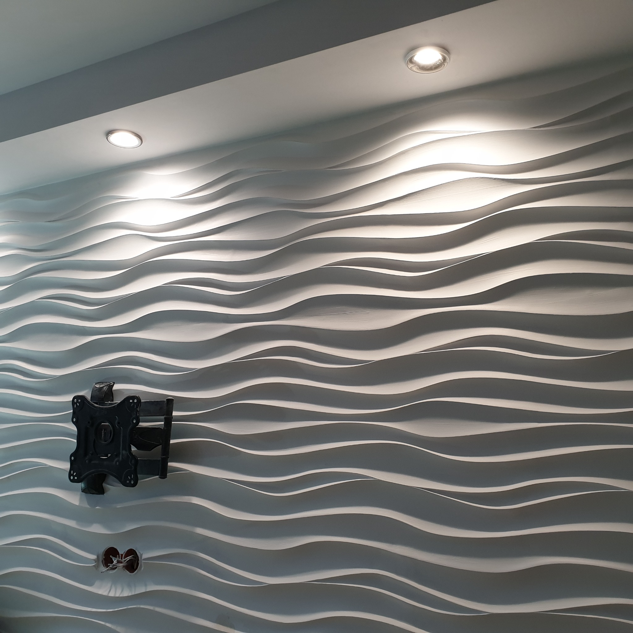 Wzorzysta, biała ściana 3D z zamontowanym uchwytem do telewizora i widocznymi otworami z przewodami elektrycznymi.