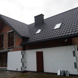 Drózd Piotr - Dom w trakcie ocieplania: widoczne białe płyty izolacyjne na ścianach zewnętrznych, surowa cegła na wyższych partiach budynku, ciemna dachówka i okna dachowe.