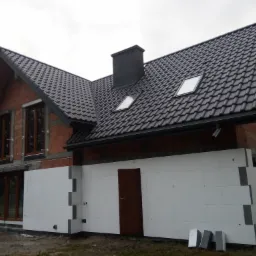 Dom w trakcie ocieplania: widoczne białe płyty izolacyjne na ścianach zewnętrznych, surowa cegła na wyższych partiach budynku, ciemna dachówka i okna dachowe.