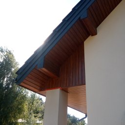 Drózd Piotr - Drewniana podbitka dachowa z ciemnymi elementami wykończeniowymi, widok z dołu na fragment elewacji budynku i zieleń w tle.