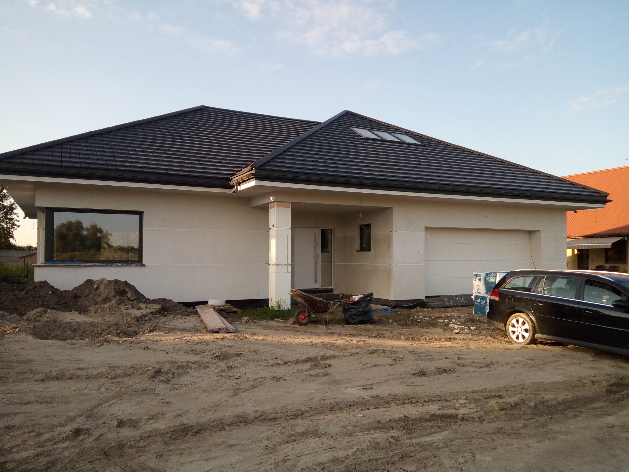 Nowoczesny dom jednorodzinny z ciemną dachówką w trakcie wykończenia, widoczny fragment elewacji, okno z odbiciem nieba, teren wokół domu częściowo uporządkowany, zaparkowany samochód osobowy.