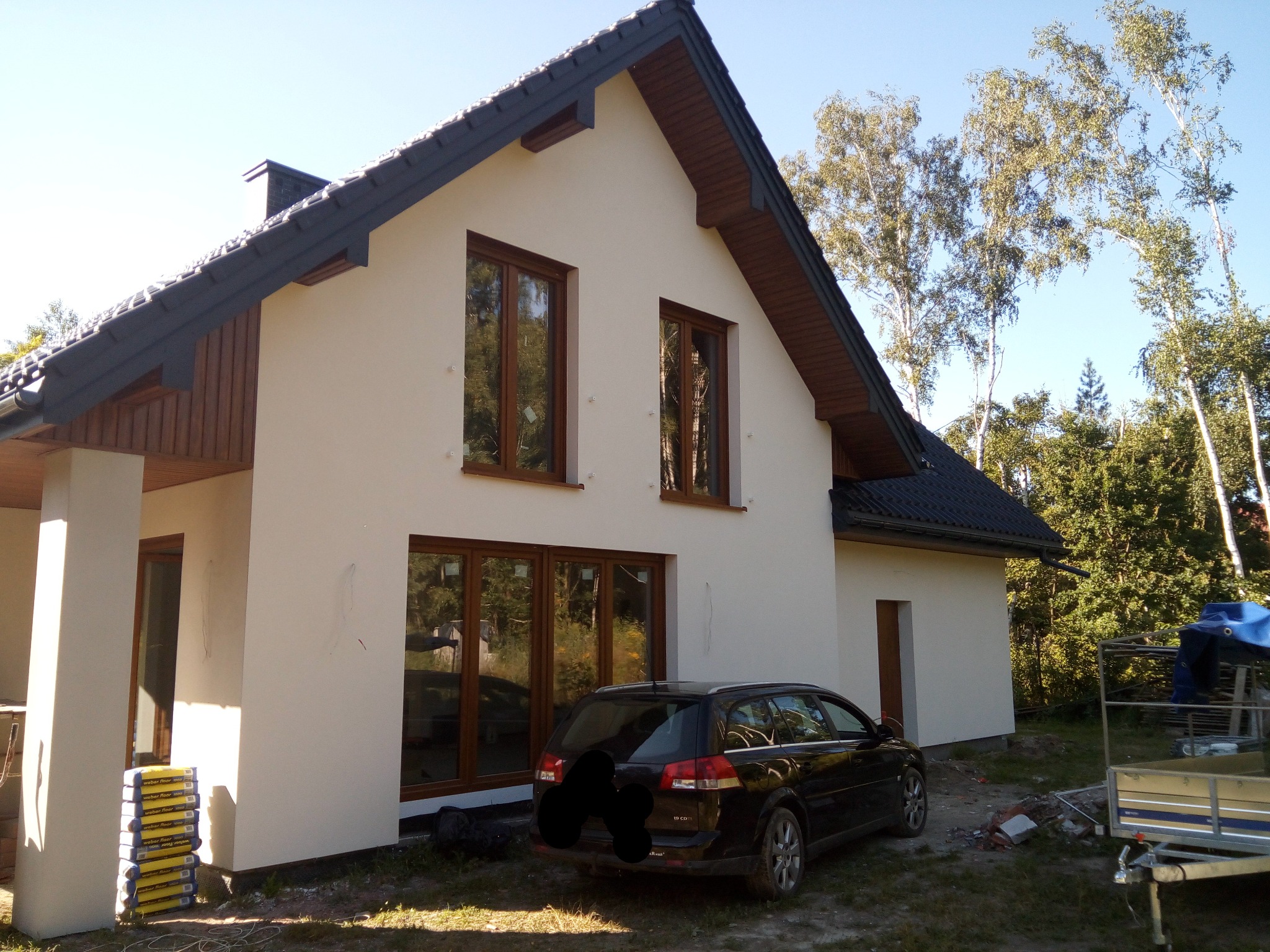 Nowo otynkowany dom jednorodzinny z brązowymi oknami i ciemnym dachem, z zaparkowanym czarnym kombi i stertą worków z zaprawą obok kolumny werandy.